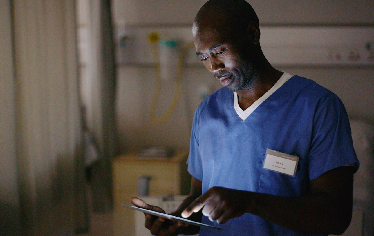 A nurse wearing scrubs in a hospital room making notes on a tablet.