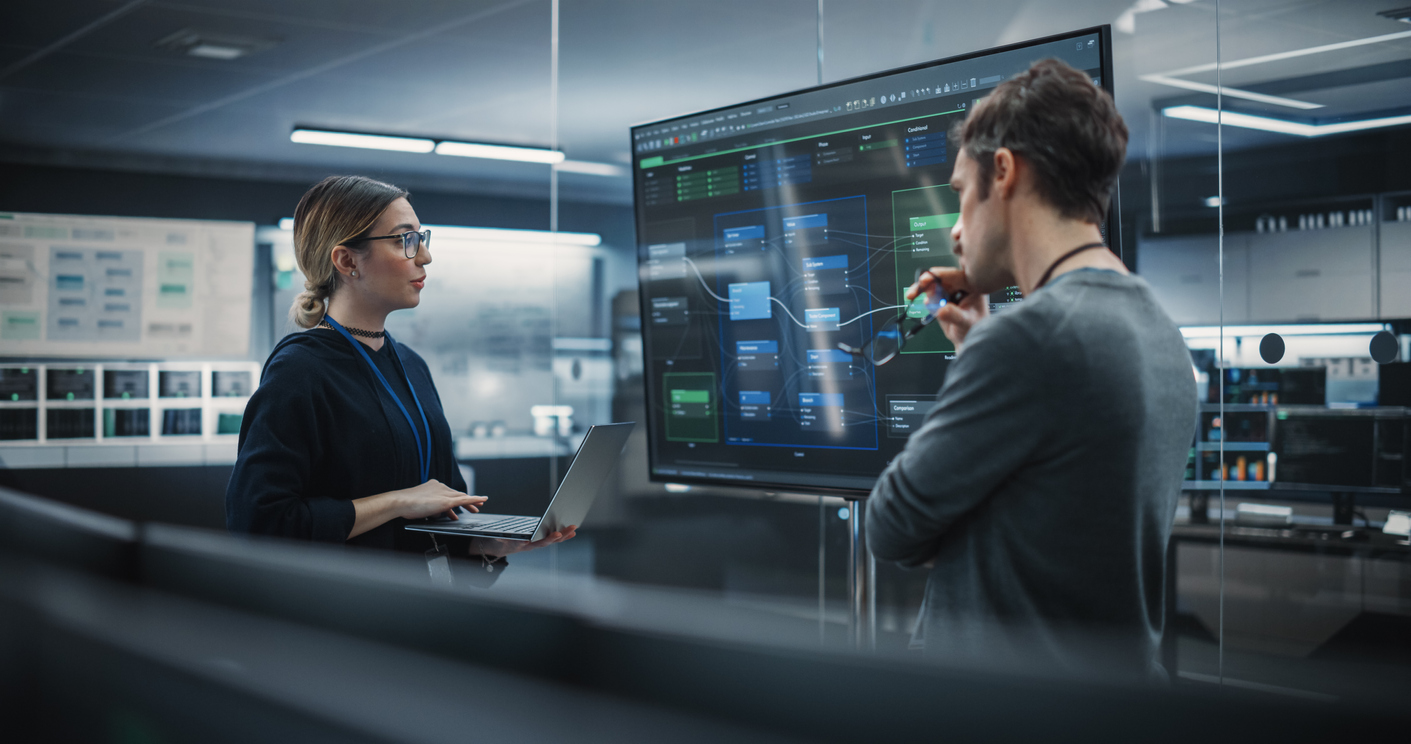 Two Cybersecurity Specialists Study a System Layout on a Large Computer Monitor.