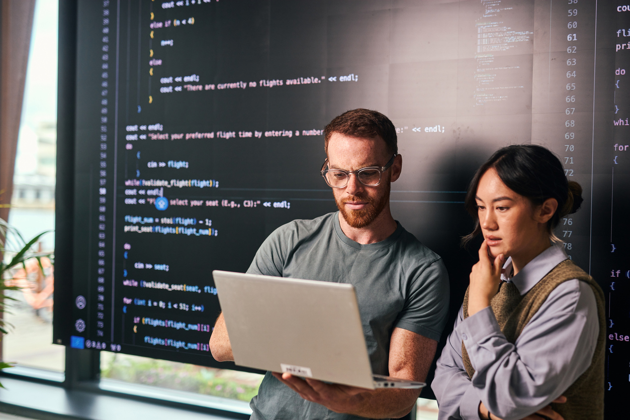 Two cybersecurity professionals consult a laptop with a smartboard behind them.